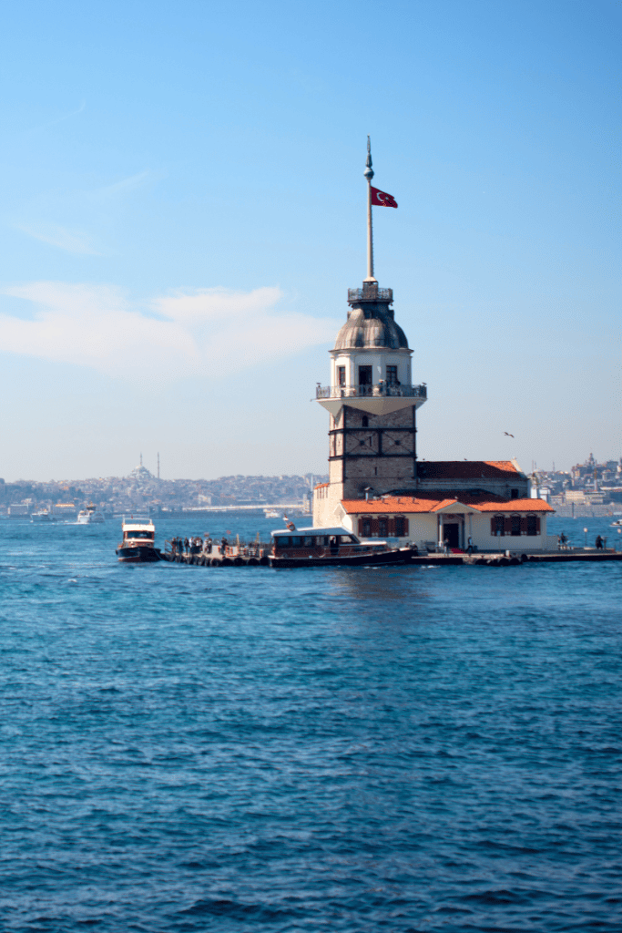 A view of the Maiden's Tower in Istanbul, standing on a small island surrounded by the sea, with a Turkish flag flying atop the structure and a clear blue sky in the background.
