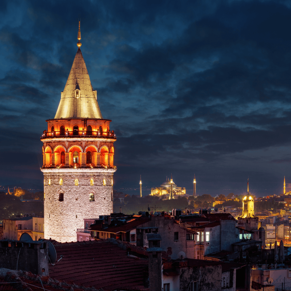 The Galata Tower illuminated at night, surrounded by city rooftops and a cloudy sky, with historical buildings in the background.